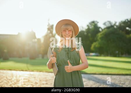 Allegra giovane bionda studentessa universitaria o universitaria con computer portatile che indossa un abito verde e cappello nel campus universitario mostrando pollici verso l'alto gesto. Felice scuola o studentessa, concetto di istruzione Foto Stock