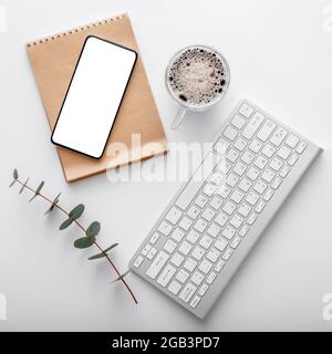 Smartphone mock-up con tastiera nota caffè tazza verde pianta su scrivania spazio di lavoro bianco. Telefono cellulare su un moderno spazio di lavoro. Vista dall'alto in piano Foto Stock