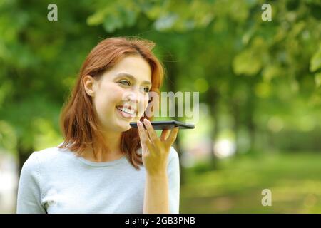 La donna felice usa il riconoscimento vocale sullo smartphone che cammina in un parco Foto Stock