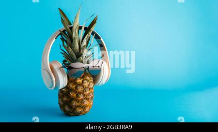 Creative divertente ananas viso indossare occhiali da sole cuffie. Ananas viso levitating ascolto di musica su sfondo estivo blu colore. Nastro lungo Foto Stock