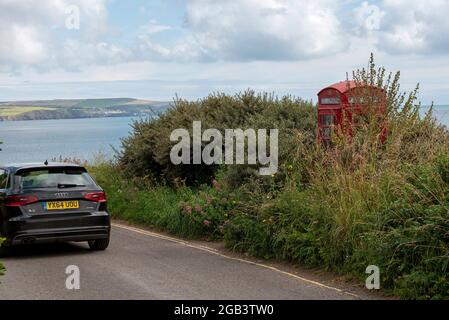 Bigbury on Sea, Devon, Inghilterra, Regno Unito. 2021. Auto che passa un telefono rosso su una strada costiera nel Devon sud, coperto di sottobosco. Foto Stock