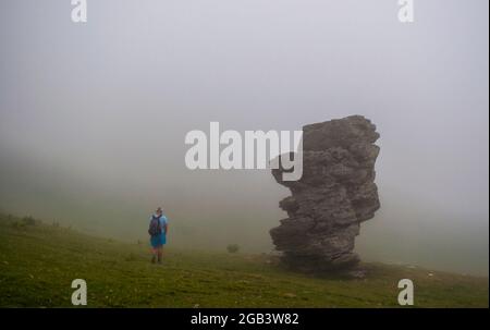 Un uomo sta camminando sulla montagna attraverso una fitta nebbia, una grande roccia è accanto a lui. Foto Stock