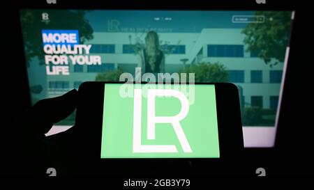 Persona che tiene il telefono mobile con il logo della società di cosmetici LR Health and Beauty Systems GmbH sullo schermo davanti alla pagina web. Mettere a fuoco il display del telefono. Foto Stock