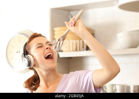 Donna felice che ascolta la musica con le cuffie che cantano utilizzando una forchetta come microfono in cucina Foto Stock