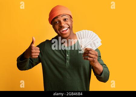 Grande profitto. Cheerful Black Man Holding Dollar Cash e mostrando Thumb Up Foto Stock