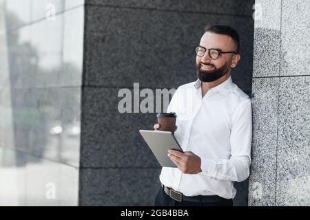Lavoro online moderno in città e tecnologia mobile. Felice lavoratore maturo in abbigliamento formale in piedi con il caffè per andare Foto Stock