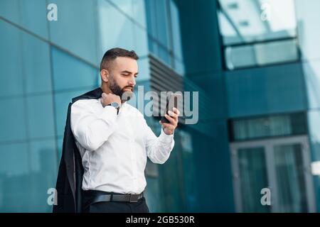 Uomo d'affari maturo ed elegante con smartphone in mano che legge un buon messaggio, in piedi vicino all'ufficio della città sulla strada, spazio libero Foto Stock