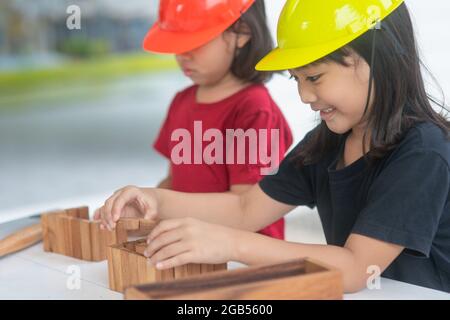 Fratelli asiatici ragazze che indossano cappelli di ingegneria costruzione Casa dal giocattolo di legno. Per imparare e migliorare lo sviluppo, piccolo architetto. Foto Stock