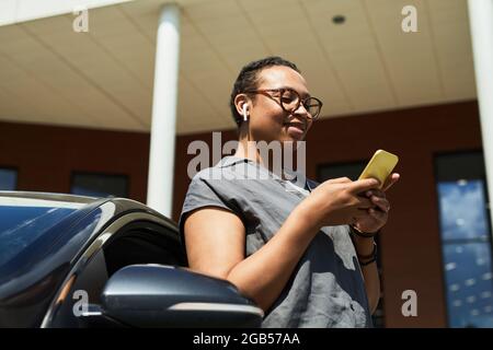 Donna africana che esce dall'auto leggendo un messaggio sul suo telefono cellulare e sorridendo Foto Stock