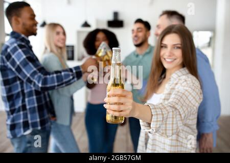 Giovane caucasica con i suoi amici multirazziali tostando con bottiglie di birra, celebrando qualcosa a casa, primo piano Foto Stock