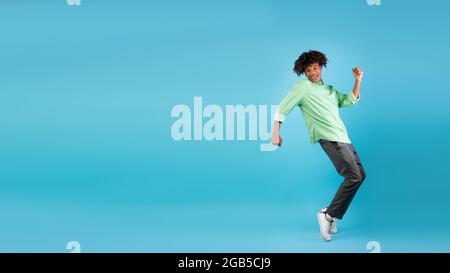 Eccitato ragazzo teen afroamericano imbrogliare, in piedi su sneakers Typtoes su sfondo blu, panorama con spazio libero Foto Stock