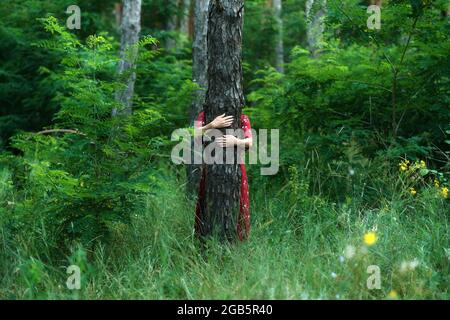 Salvare e piantare alberi, salvare la Terra e la natura, stare per gli alberi, salvare le foreste e guarire il clima. Donna in un vestito rosso che cammina nella foresta, abbracciando gli alberi Foto Stock