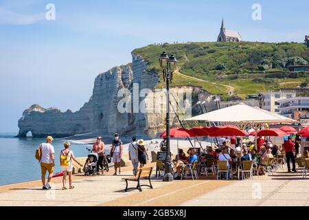 La gente che beve un drink in un bar sulla spiaggia sul lungomare di Etretat, in Francia, con la cappella di Notre-Dame de la Garde che si affaccia sulla scogliera di Amont e il suo arco. Foto Stock
