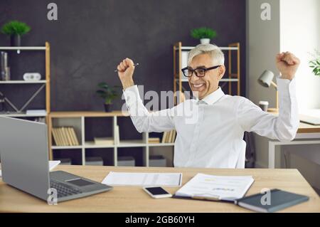 Uomo felice seduto alla scrivania dell'ufficio, guardando lo schermo del computer portatile e sorridendo Foto Stock