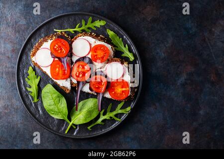 Toast sani con formaggio cremoso, pomodori ciliegini e spinaci su piatto nero. Bruschetta con formaggio cremoso ed erbe fresche su fondo scuro. Copia Foto Stock