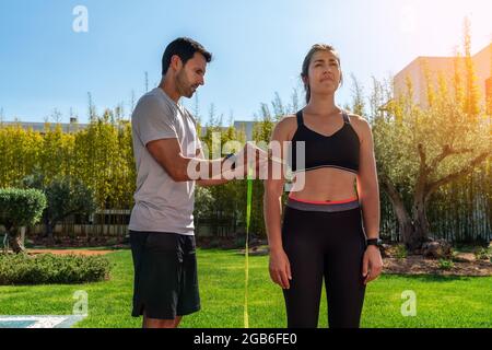 Un uomo europeo, personal trainer, misura la mano del cliente, l'anca per il progresso nella formazione, per la perdita di peso. In estate, in giardino con piscina. Foto Stock