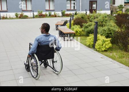 Vista posteriore di un disabile afro-americano che si muove in avanti in sedia a rotelle mentre si cammina per la strada Foto Stock