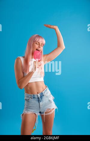 Donna vivace con capelli rosa che si diverte, tenendo il pettine per capelli e cantando in esso mentre si trova isolato su sfondo blu studio Foto Stock