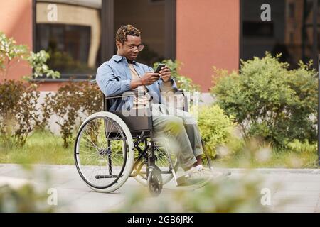 Contenuto uomo afroamericano handicappato seduto in sedia a rotelle sulla strada e leggendo un messaggio al telefono mentre chattava online Foto Stock