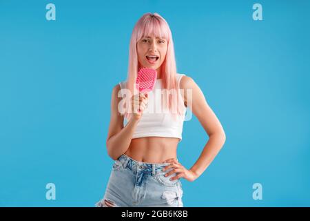 Donna gioiosa con capelli rosa che tengono il pettine per capelli e che canta in esso mentre si trova isolato su sfondo blu studio Foto Stock