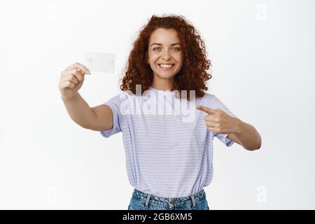 Immagine di bella ragazza curly che punta e tiene la carta di credito, diventare cliente di banca, annunciando sconti, in piedi su sfondo bianco Foto Stock