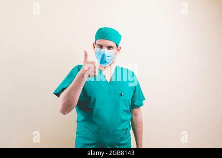 Ritratto giovane medico con pollice in tuta verde chirurgo e maschera respiratoria protettiva sullo sfondo giallo isolato Foto Stock
