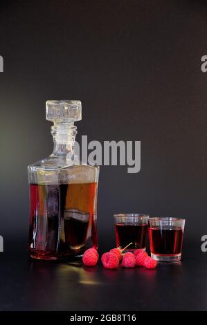 Liquore fatto in casa di lamponi in due bicchieri e una bottiglia, bacche mature nelle vicinanze su sfondo nero. Primo piano. Foto Stock