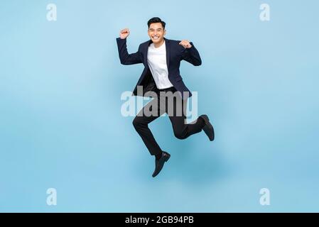 Ritratto a tutta lunghezza di un bell'uomo asiatico estatico che salta e alza i pugni sullo sfondo isolato di uno studio azzurro Foto Stock