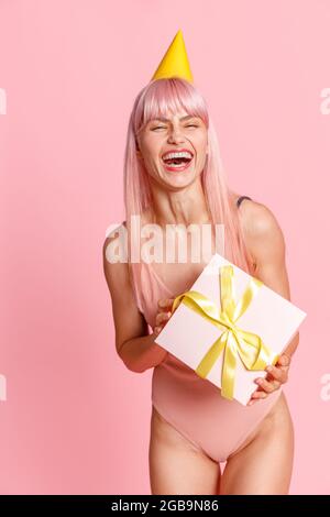 Giovane donna eccitata con i capelli rosa che indossano il costume da bagno e cappello da festa che tiene la scatola del regalo carina, in piedi isolato su sfondo rosa Foto Stock