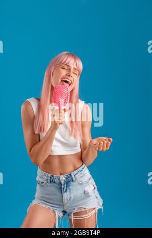 Giovane donna divertente con capelli rosa che tengono il pettine per capelli e cantare in esso come una stella pop mentre si trova isolato su sfondo blu studio Foto Stock