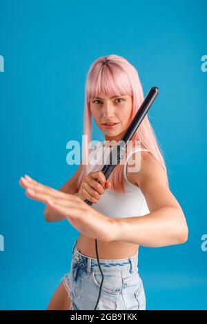 Giocosa giovane donna con i capelli rosa guardando la macchina fotografica, facendo arti marziali posa e tenendo arricciante ferro, in posa isolato su sfondo blu studio Foto Stock