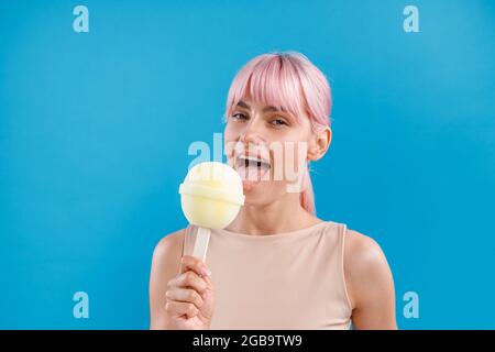 Ritratto di donna carina con i capelli rosa guardando la macchina fotografica e tenendo un grande lecca lecca gelato davanti a lei, in posa con bocca aperta isolata Foto Stock