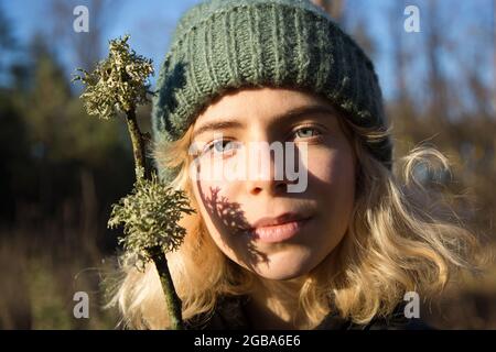 ritratto di una ragazza adolescente di 17 anni in un cappello a maglia. Giornata di sole, passeggiata nella foresta, detox digitale. Tiene il bastone con il lichen in mano davanti a f Foto Stock