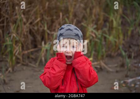 ritratto di carino bambino in giacca rossa con un'emozione luminosa sul viso. guarda in cornice, mani schiacciate alle guance, bocca aperta con rabbia o Foto Stock