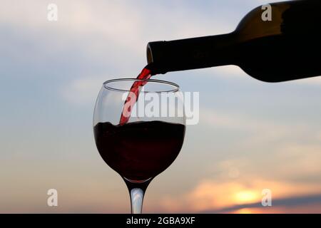 Vino rosso che si versa da una bottiglia nel bicchiere sullo sfondo del tramonto. Concetto di celebrazione, festa estiva al resort, cena romantica all'aperto Foto Stock