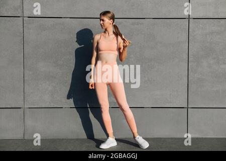 Giovane donna fitness con un bel corpo, in abiti sportivi rosa, esercizio all'aperto, sullo sfondo di una parete grigia urbana, modello femminile in Foto Stock