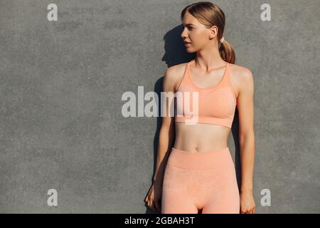 Giovane donna fitness con un bel corpo, in abiti sportivi rosa, esercizio all'aperto, sullo sfondo di una parete grigia urbana, modello femminile in Foto Stock
