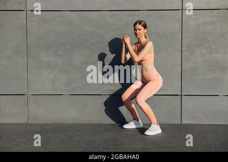 Vista laterale scatto di giovane donna in forma che fa squats, l'esercizio sulle natiche all'aperto, l'allenamento sulla parete di cemento urbano sfondo, fitness femminile esercizio outd Foto Stock