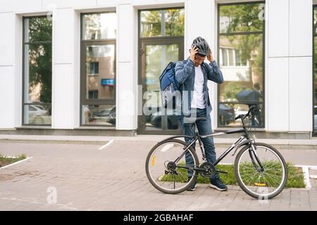 Cheerful Courier maschio uscire dall'appartamento del cliente, seduto in bicicletta e uscire alla prossima consegna. Foto Stock