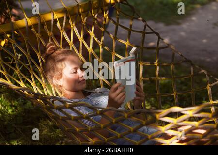 Giovane studentessa sorridente a leggere un libro mentre si steso su un'amaca in giardino. Donna che si rilassa nel libro di lettura amaca. Torna all'università, all'università Foto Stock