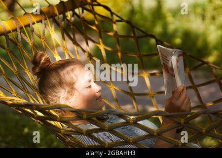 Giovane studentessa sorridente a leggere un libro mentre si steso su un'amaca in giardino. Donna che si rilassa nel libro di lettura amaca. Torna all'università, all'università Foto Stock