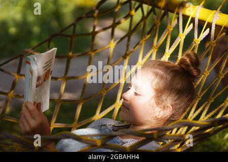 Giovane studentessa sorridente a leggere un libro mentre si steso su un'amaca in giardino. Donna che si rilassa nel libro di lettura amaca. Torna all'università, all'università Foto Stock