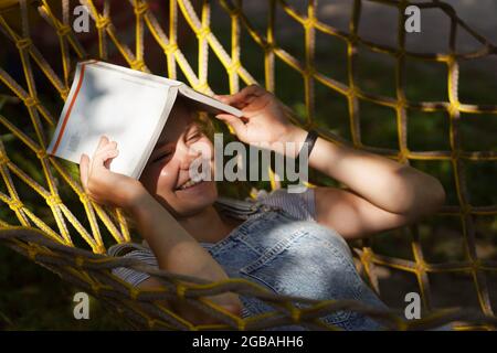 Giovane studentessa sorridente a leggere un libro mentre si steso su un'amaca in giardino. Donna che si rilassa nel libro di lettura amaca. Torna all'università, all'università Foto Stock