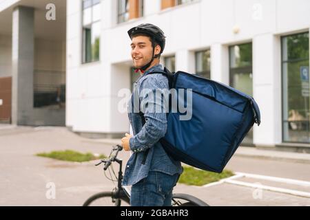 Vista posteriore di un bel ragazzo sorridente in casco protettivo che posa in piedi vicino alla bicicletta in via della città sullo sfondo dell'edificio degli uffici Foto Stock