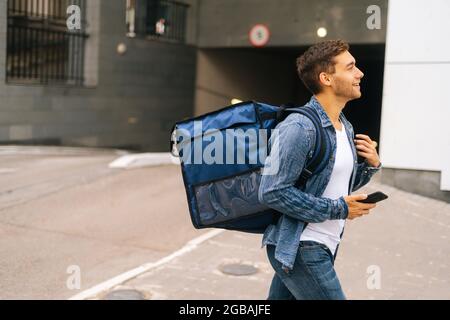 Vista laterale del bel corriere maschile con termo zaino camminare in città strada tenendo il telefono cellulare è mano. Foto Stock