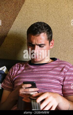 Uomo serio giovane 20s che tiene piccola chitarra - ukulele e smartphone mobile. Sorprendo ragazzo caucasico marrone con capelli rossi all'interno. Freelance o uomo d'affari. Foto Stock