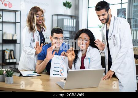 Quattro diversi medici di genetica multirazziale scienziati in uniformi che ondolano al loro collega durante la video conferenza online, mentre lavorando sulla ricerca congiunta di formula di DNA strutturale e farmaci Foto Stock