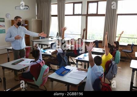 Gruppo di studenti diversi che indossano maschere di protezione sollevando le mani nella classe a scuola Foto Stock