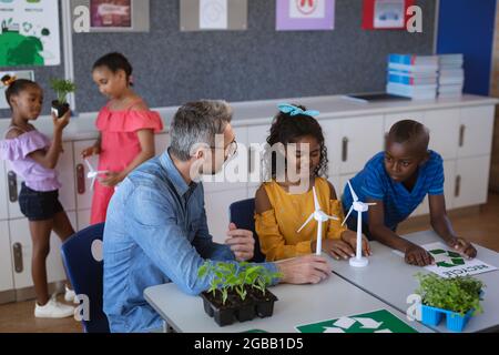 Insegnante di sesso maschile caucasico che tiene il modello di mulino a vento insegnando ragazzo e ragazza in classe ambiente a scuola Foto Stock