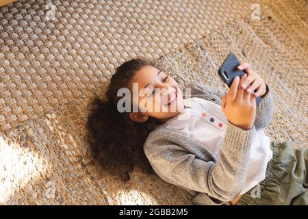 Sorridente ragazza da corsa mista che si stesa sul tappeto utilizzando uno smartphone Foto Stock